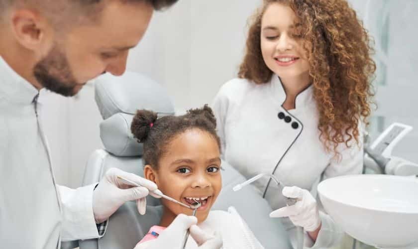 Pediatric dentist and assistant examining young girl's teeth during dental checkup.