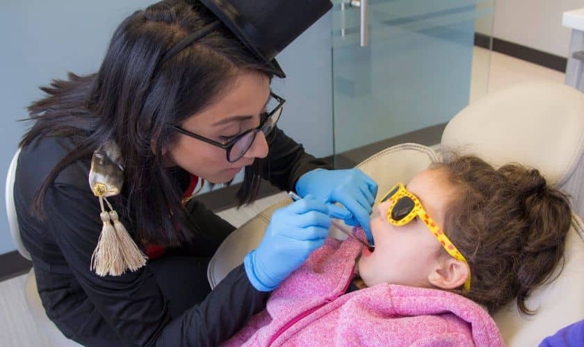 Pediatric dentist in blue gloves examining young patient's teeth during festive dental visit.