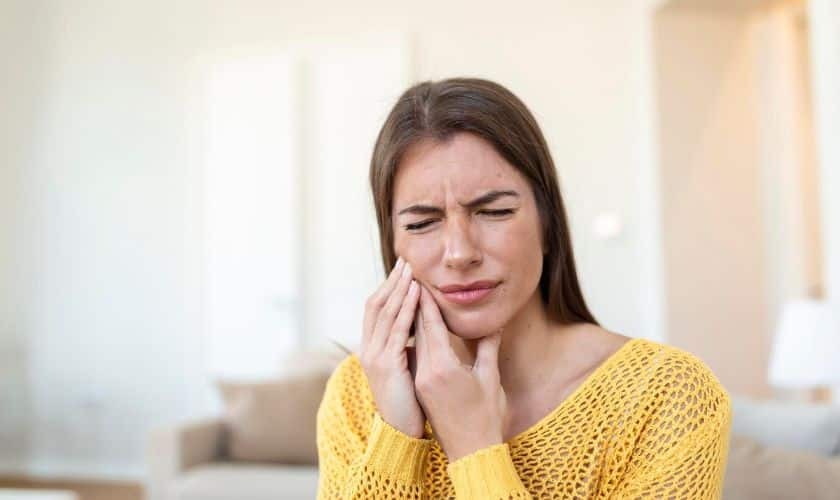 Woman in yellow sweater holding her cheek in pain, indicating dental discomfort or toothache.