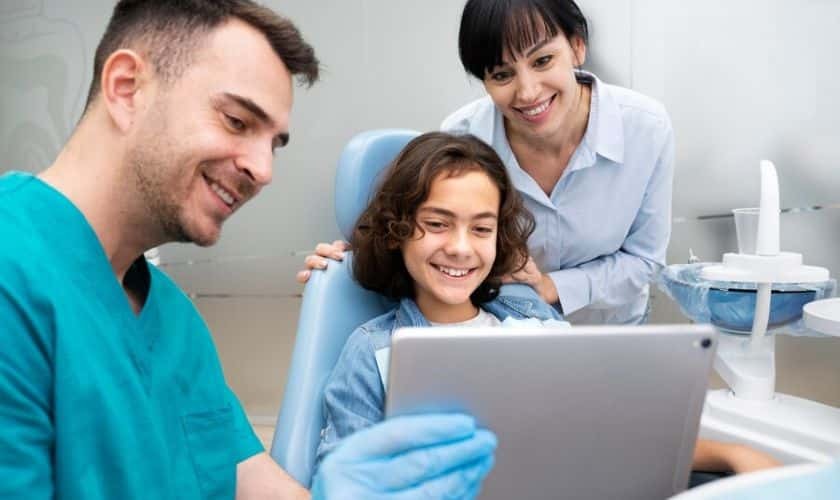 Pediatric dentist and assistant showing young patient dental results on tablet in treatment room.