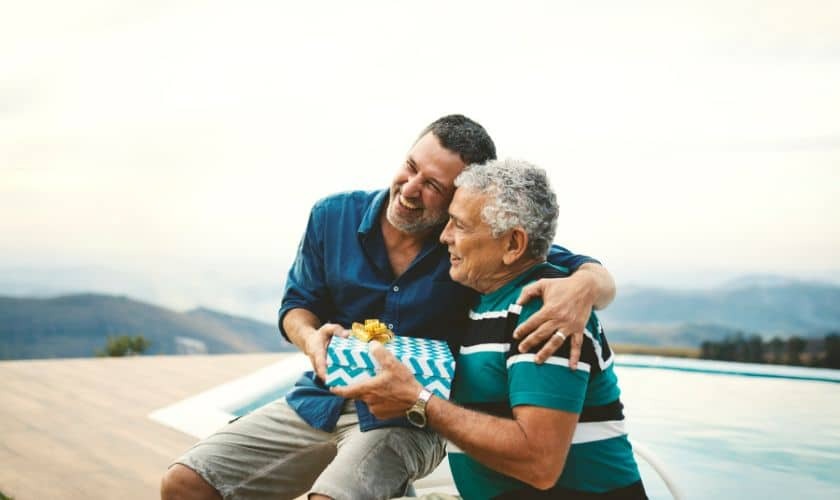 Adult son giving wrapped gift to father outdoors with mountain view backdrop.