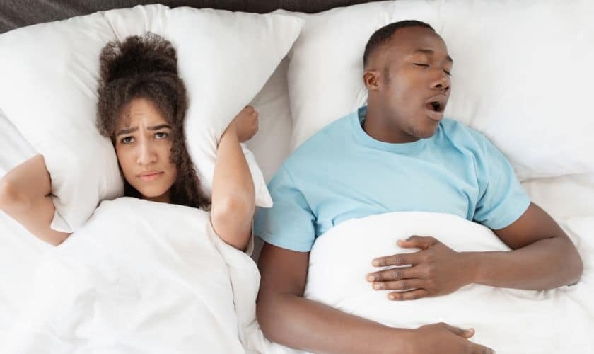 Couple in bed where man snores while woman looks frustrated, illustrating sleep apnea concerns.