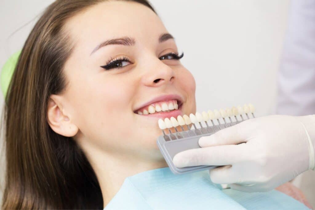 Woman smiling while dentist holds tooth shade guide for cosmetic dental treatment planning.