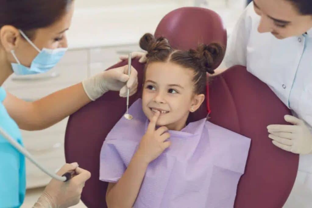 Young patient smiling in dental chair during pediatric examination with dentist and assistant.