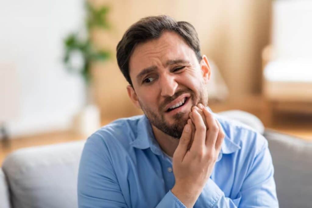 Man in blue shirt holding cheek in pain, experiencing dental discomfort.