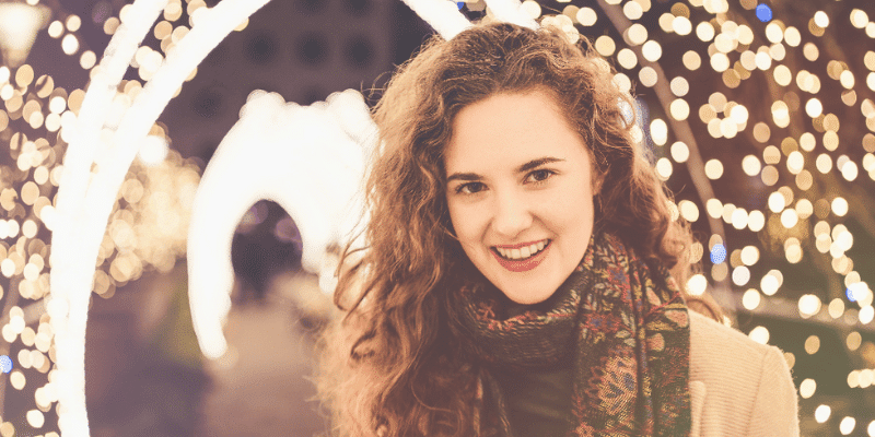 Smiling woman with curly hair enjoying holiday lights during festive season.