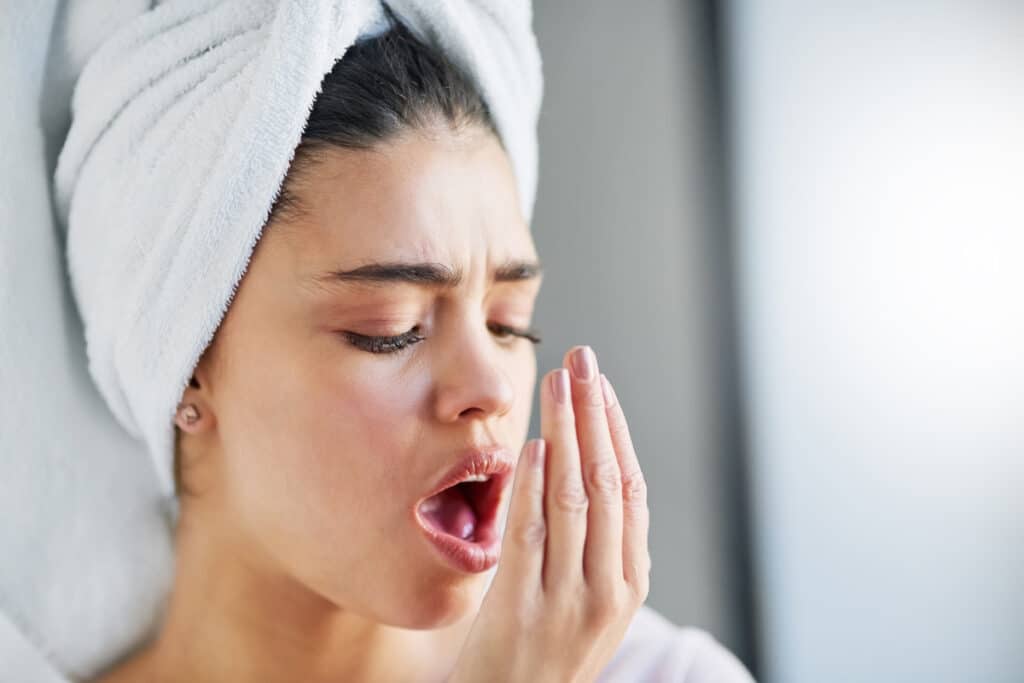 Woman with towel wrapped around head checking her breath with hand near mouth.