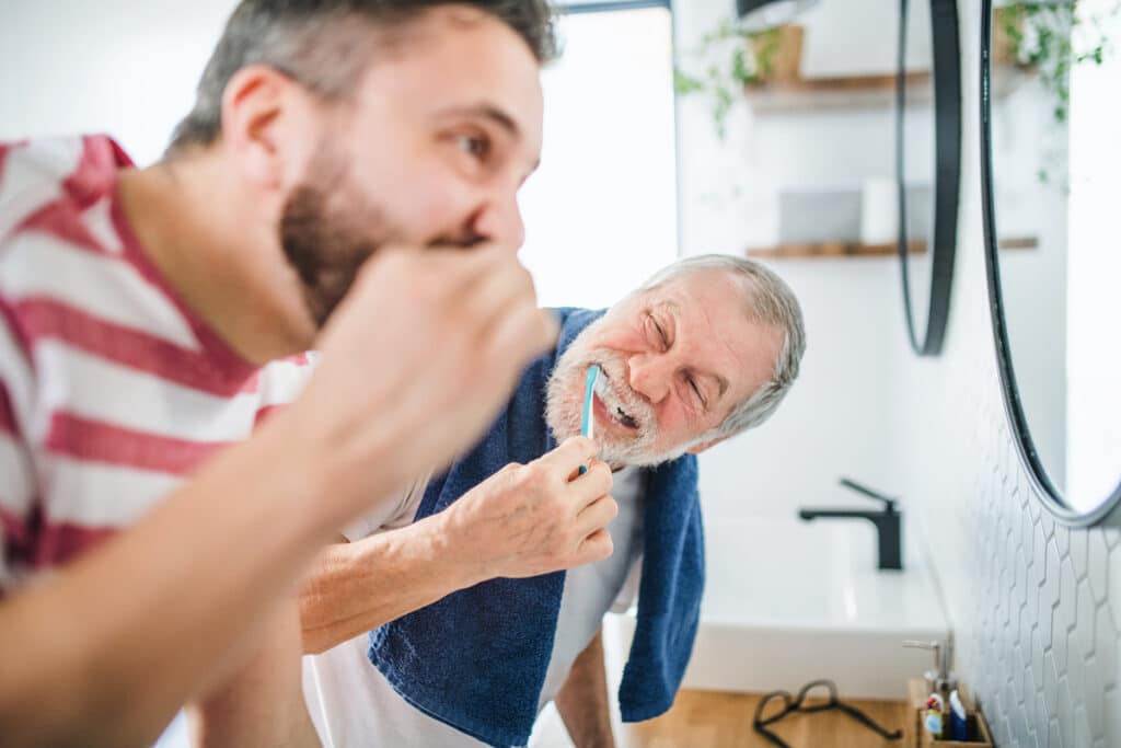 Older man brushing teeth while younger man watches in modern bathroom mirror.