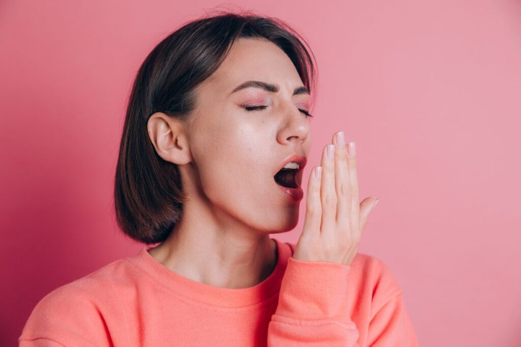 Woman demonstrating bad breath concern by cupping hand near mouth against pink background.