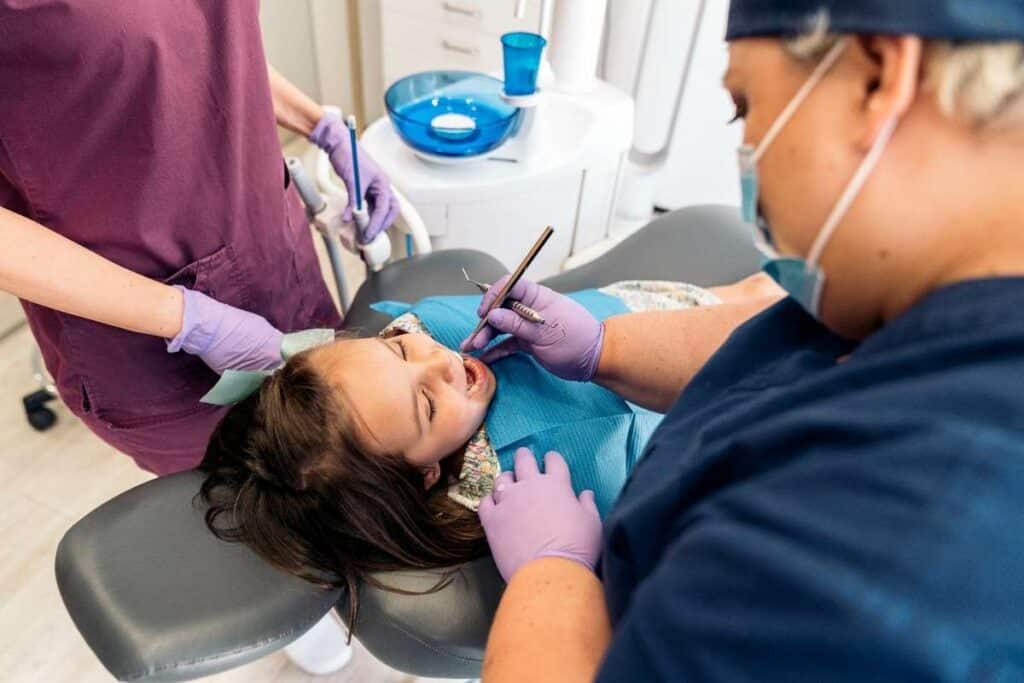 Pediatric dental examination showing dentist and assistant treating young patient in dental chair.