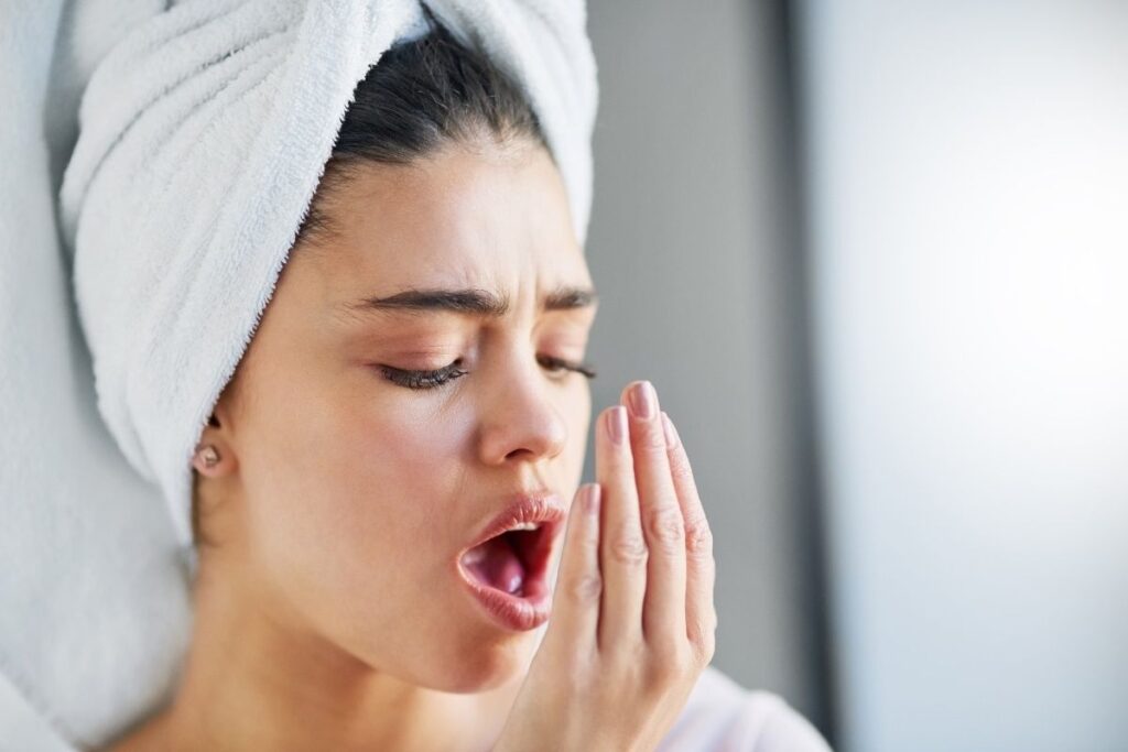 Woman with towel wrapped around head checking breath odor with hand near mouth.