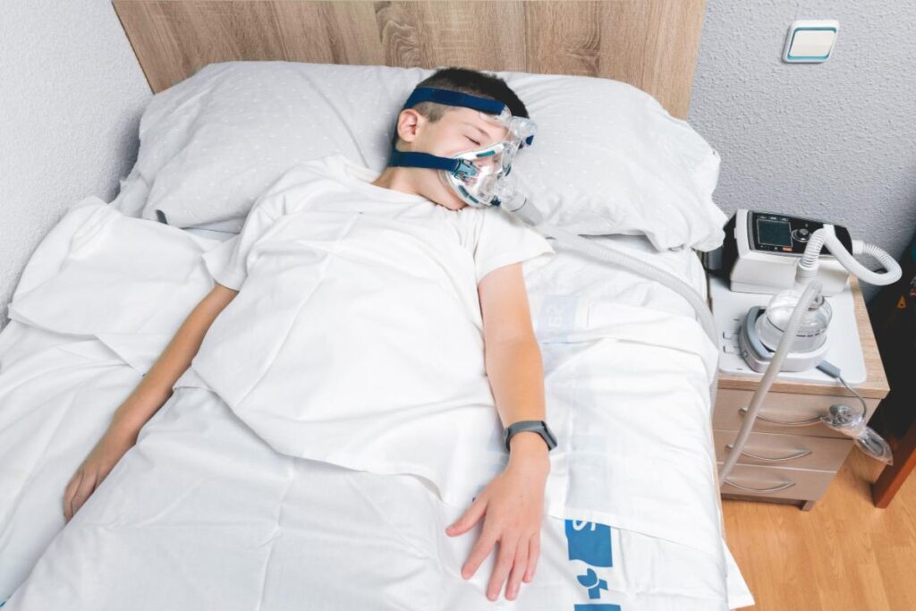 Child wearing CPAP mask sleeping in bed with sleep apnea monitoring equipment.